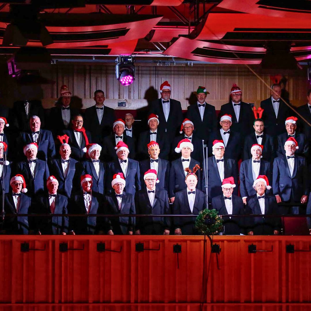 A photo of lots of male choir members singing. They are on a stage in a large hall in formal wear, except for Christmas themed hats on their heads