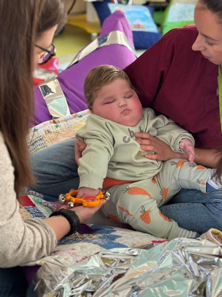 Diane helps baby Dewi shake orange bells while Gemma holds him