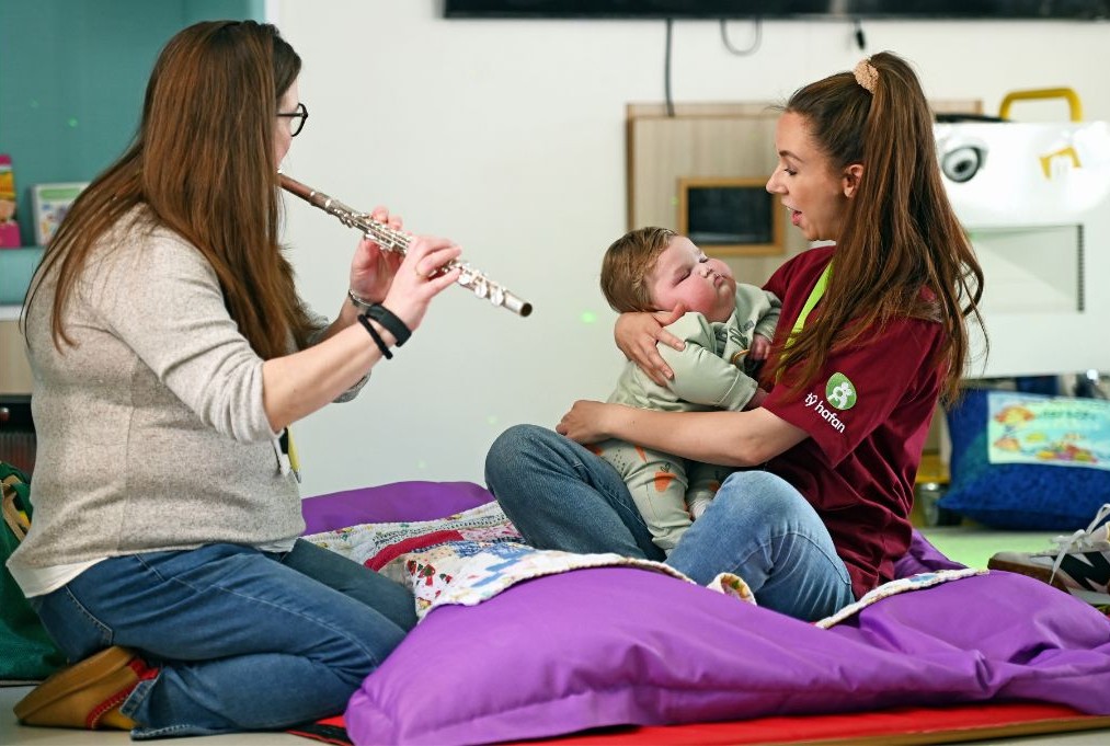 Diane plays the flute to baby Dewi, while Gemma holds him