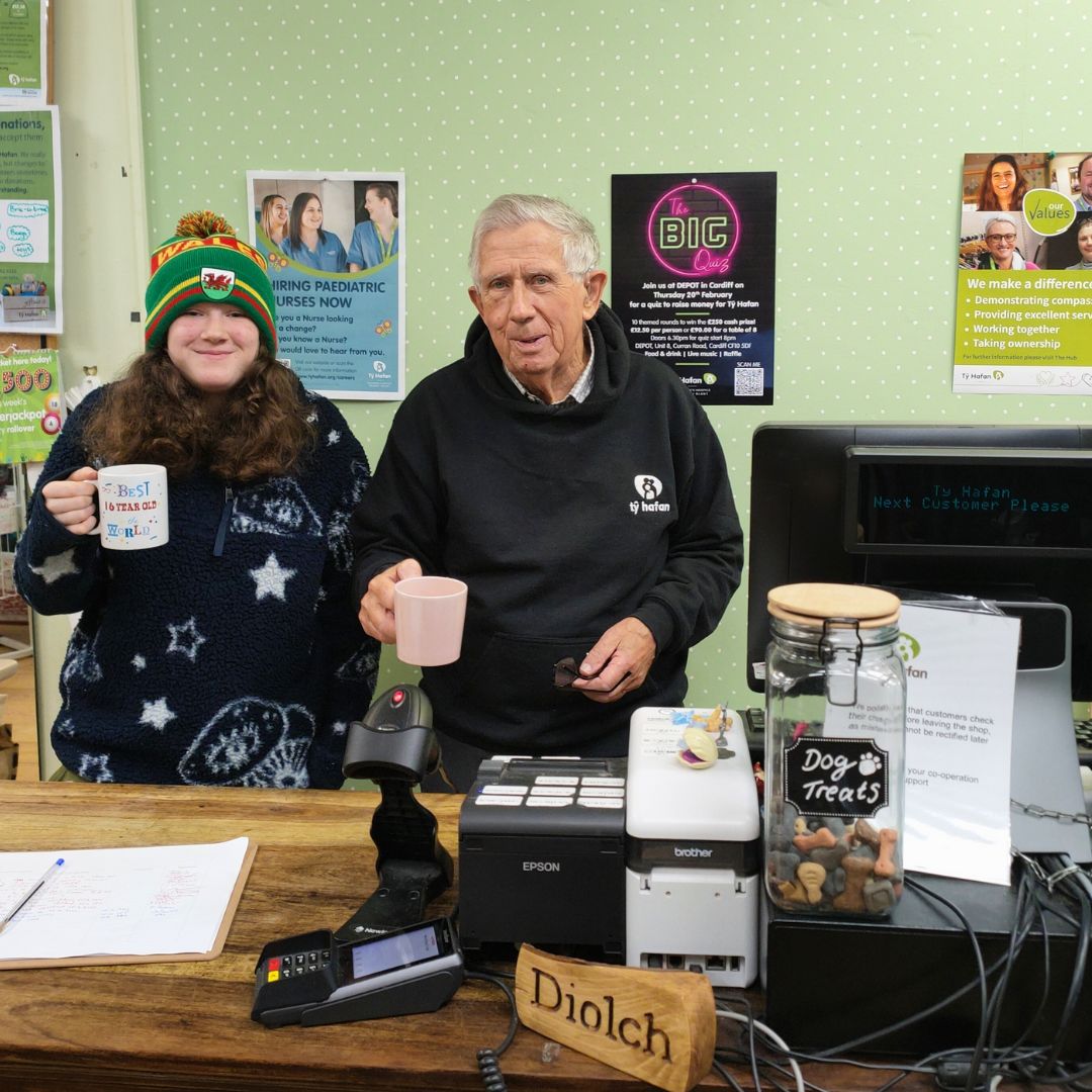 A photo of Bill and Mae behind the till at a Ty hafan charity shop. They are both holding cups of tea