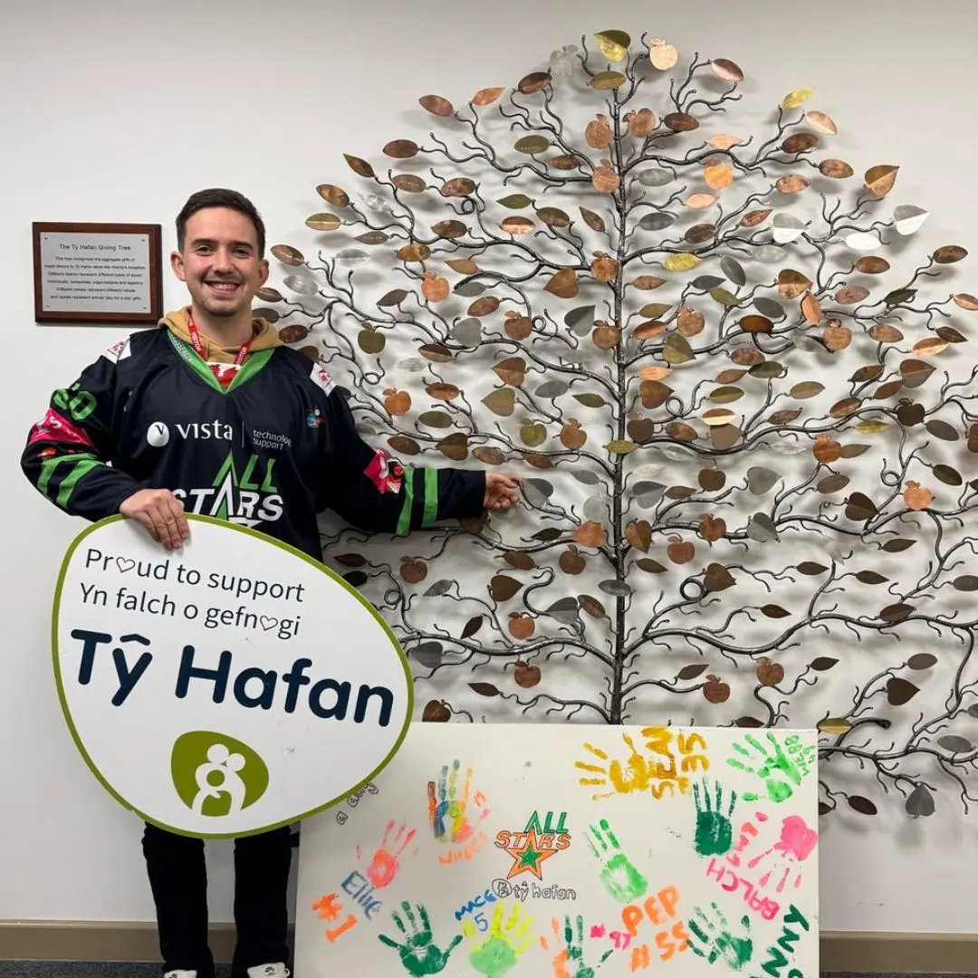 A photo of Tŷ Hafan's ice hockey team captain Scott Smith-Myers, holding a "proud to support Tŷ Hafan" sign next to our metal giving tree