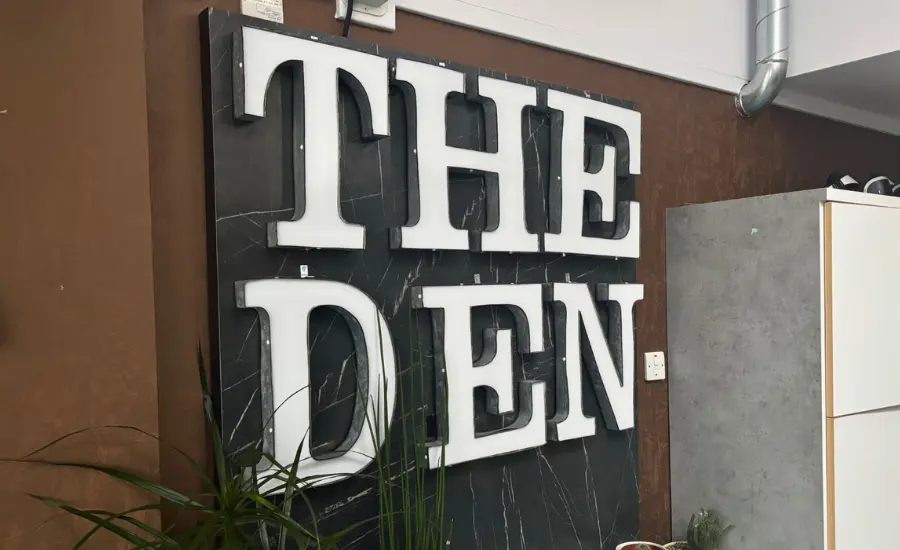 A photo of a sign in The Den. It's white text on a black background, mounted on a brown wall. It reads "The Den" and lights up