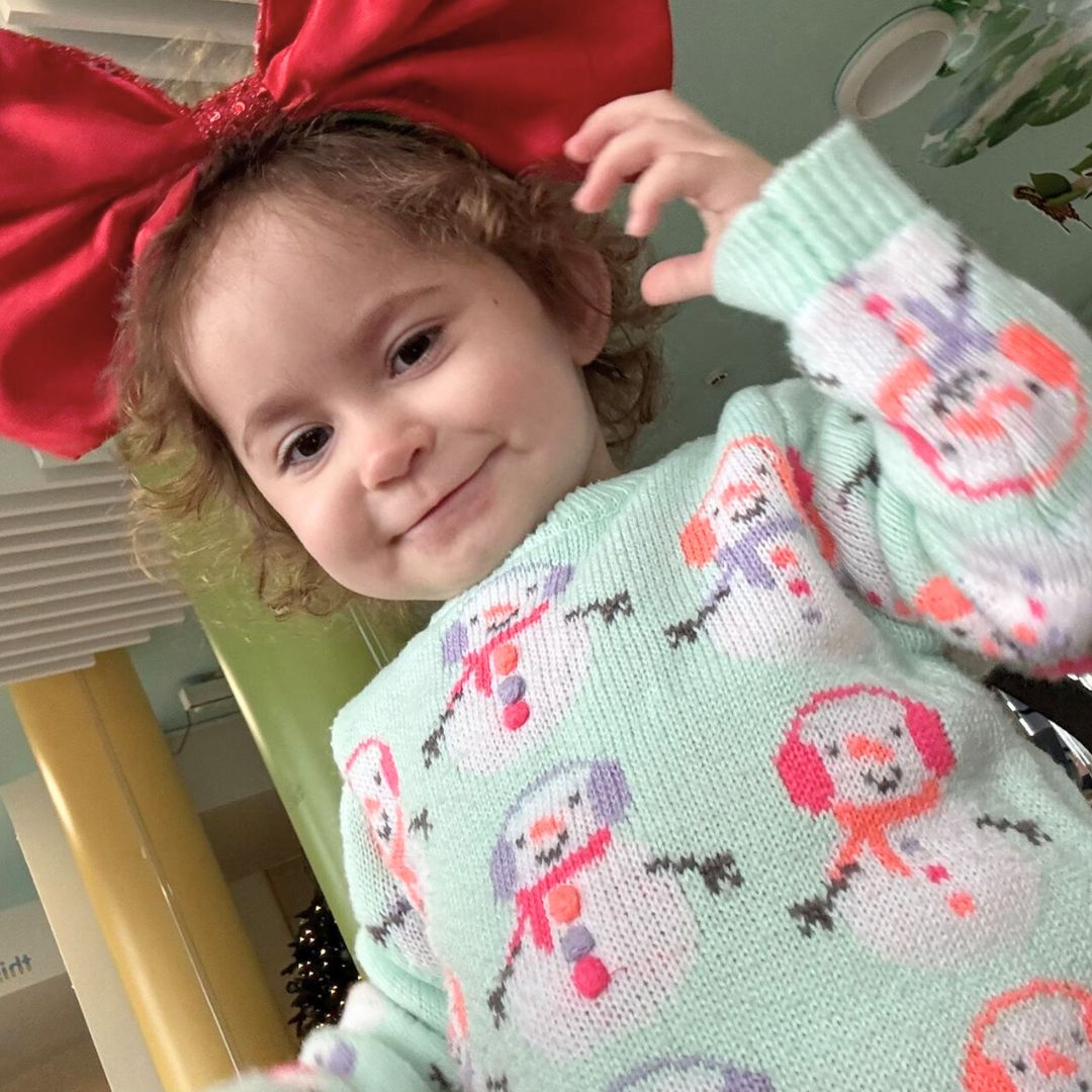 A photo of a little girl, Ivy-Mai, looking down at the camera. She has a blue bumper with snowmen on it and has put a giant red bow in her hair