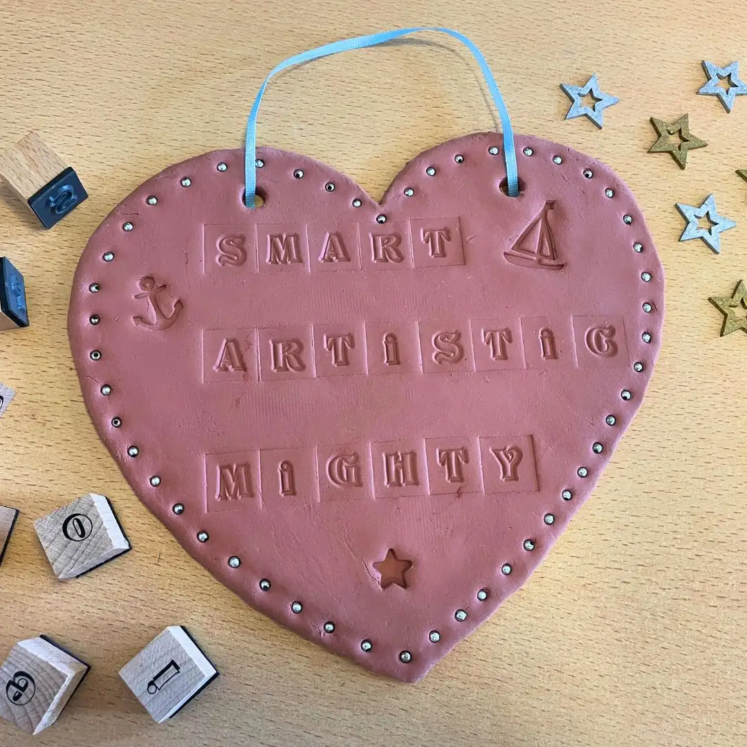 A photo of a clay heart, which has an acrostic poem stamped into it. The person's name is "Sam", and the words read "Smart, Artistic, Mighty"